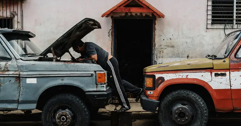 Man working on car engine outdoors, examining under the hood of classic trucks parked closely together.