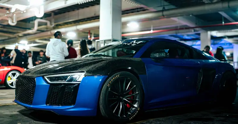 Sleek blue supercar with carbon fiber detailing at a car meet in a dimly lit garage.