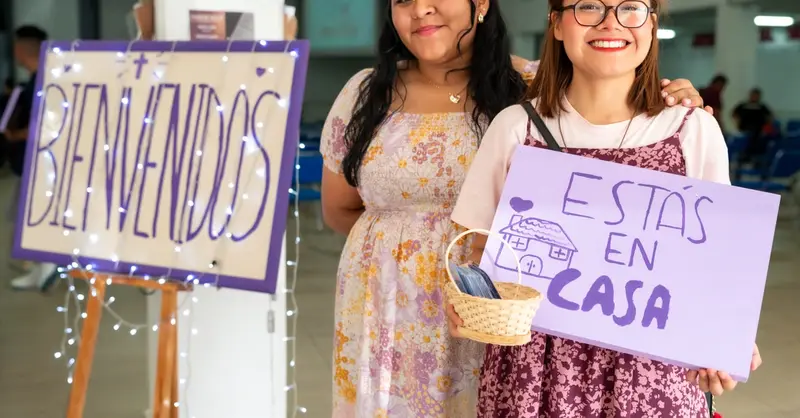 Two women warmly greet visitors at a community event in Ciudad de México with signs saying 'Bienvenidos' and 'Estás en Casa'.