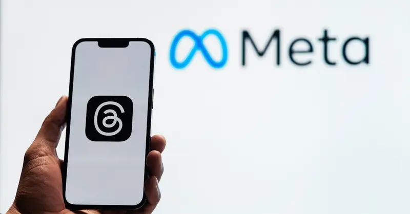 A hand holding a smartphone showing the Threads app with Meta logo in the background.