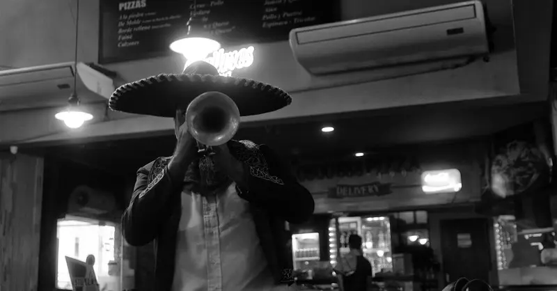 Black and white photo of a mariachi musician in a sombrero playing trumpet indoors.