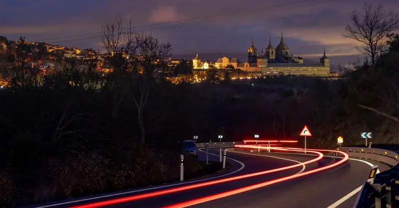Night view of El Escorial with light trails on winding road, capturing a dramatic cityscape scene.