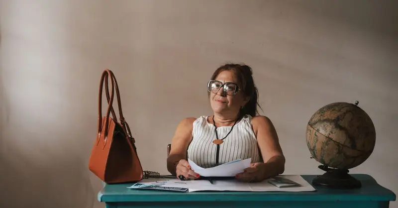 An adult woman sits at a desk with a globe, papers, and a handbag, exuding a professional appearance.