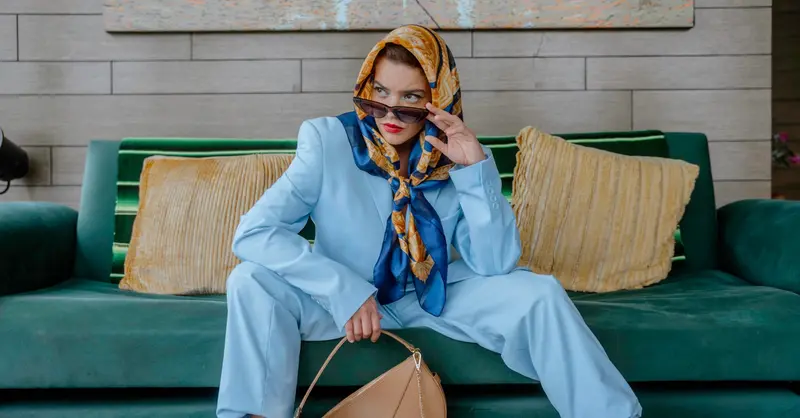 Elegant woman in a blue suit and scarf sitting stylishly on a sofa inside a living room.