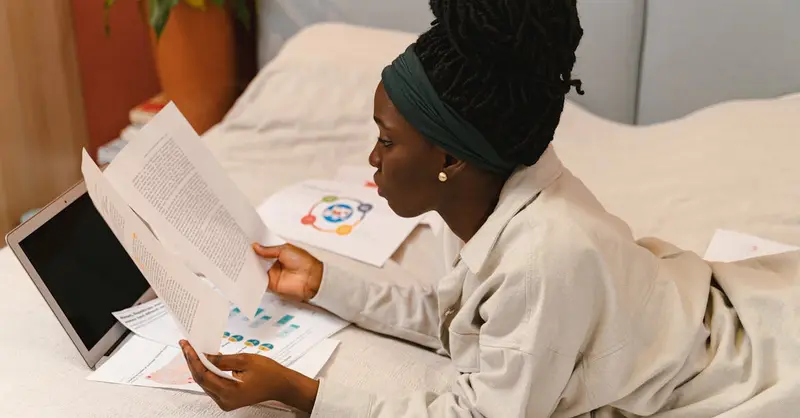 Young woman lying on bed, reading papers with a laptop nearby, focused and working indoors.