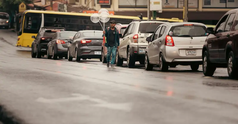 A street vendor in Alajuela, Costa Rica, selling balloons amidst traffic. Captured on a rainy day.