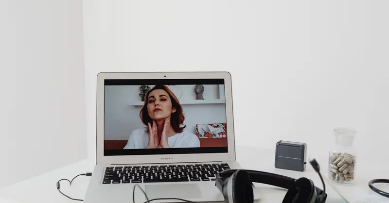 A woman conducting a telemedicine session visible on a laptop screen with headphones nearby.