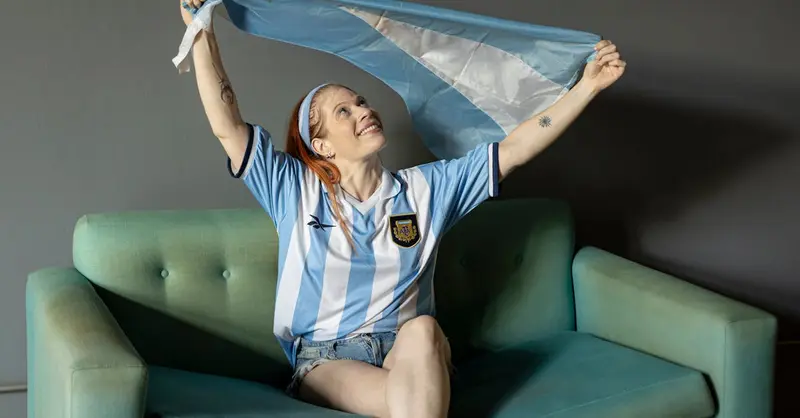 An Argentine woman cheerfully waves a flag indoors, celebrating a victorious moment.