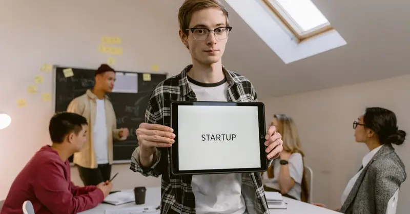 A young man in an office holding a tablet with 'STARTUP' during a meeting.