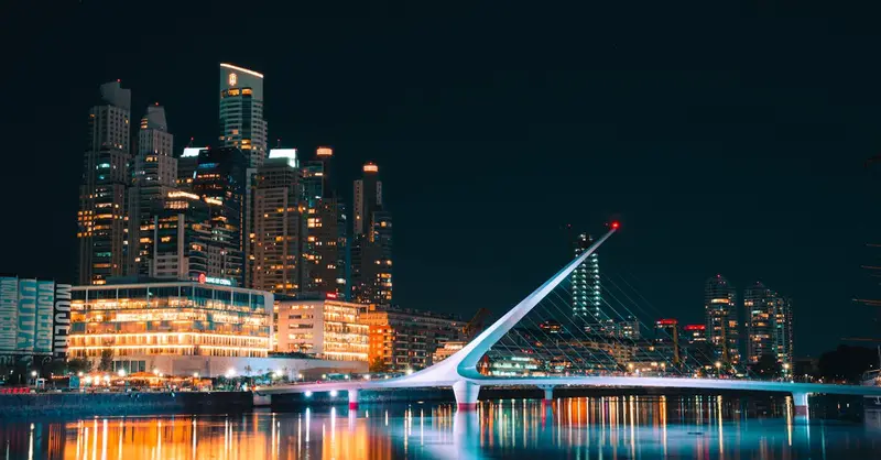 Modern architectural view of Puente de la Mujer and Buenos Aires skyline reflected in water at night.