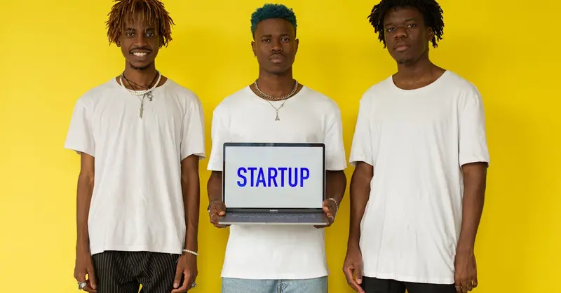 Three men holding a laptop displaying 'STARTUP' against a bold yellow background.