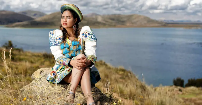 A Bolivian woman in traditional attire sitting near Lake Titicaca under a bright sky.