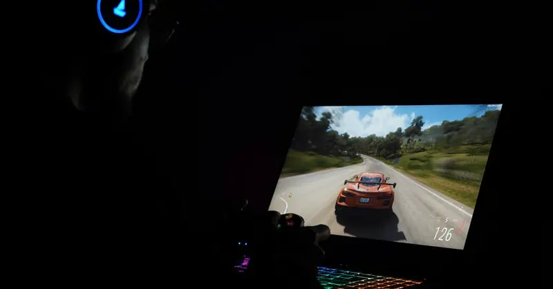 A gamer using a laptop and headphones to play a car racing game in a dark setting.