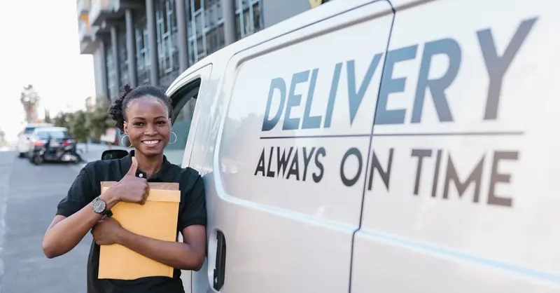 A cheerful delivery woman standing by a van with a parcel, ready for delivery.