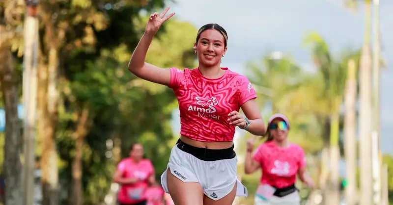 Energetic woman running outdoors in pink event shirt, smiling and flashing a peace sign.