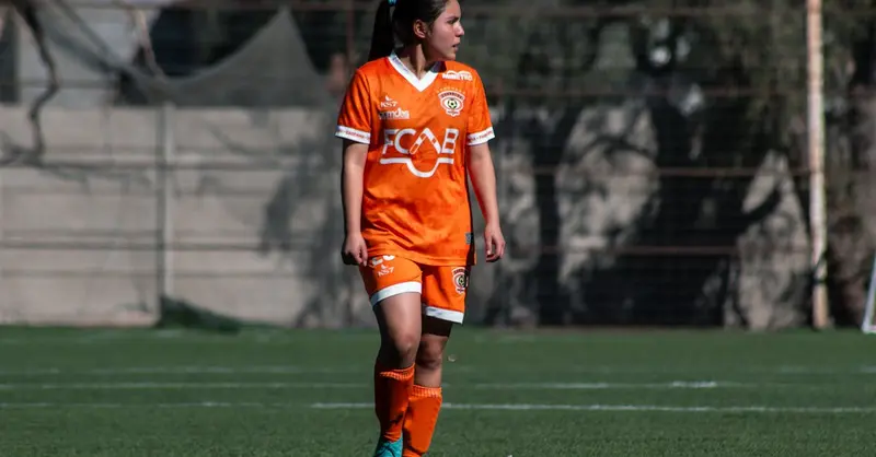 A female soccer player in orange uniform during a match on an outdoor field in Chile.