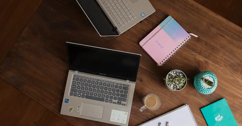 Top view of a wooden desk with laptops, notebooks, coffee cup, and potted plant. Ideal for office or study themes.