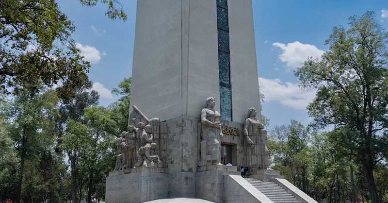 A towering stone monument with sculptures, surrounded by trees in a city park, under a bright blue sky.