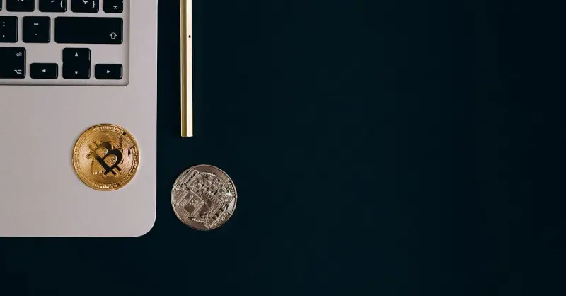 Close-up view of Bitcoin and Ethereum coins next to a laptop and pen on a black surface.