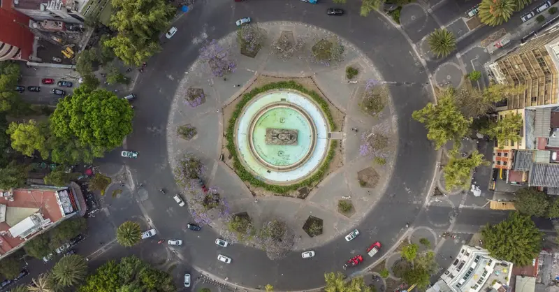 Drone shot of a scenic roundabout in Mexico City, surrounded by lush green trees and urban landscape.