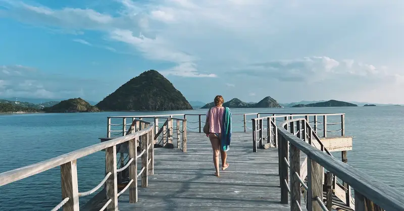 A person walks on a scenic pier in Labuan Bajo, Indonesia, with lush islands in view.