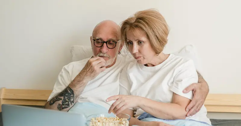Senior couple bonding over movie night with popcorn and laptop in cozy home setting.