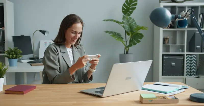 A woman in a home office using a smartphone while working on a laptop.