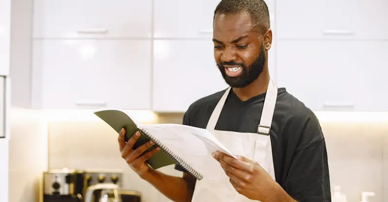 A chef wearing an apron looks puzzled while reading a recipe in a contemporary kitchen.