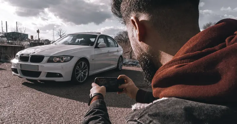 A man takes a picture of a white luxury car using a smartphone outdoors under a cloudy sky.