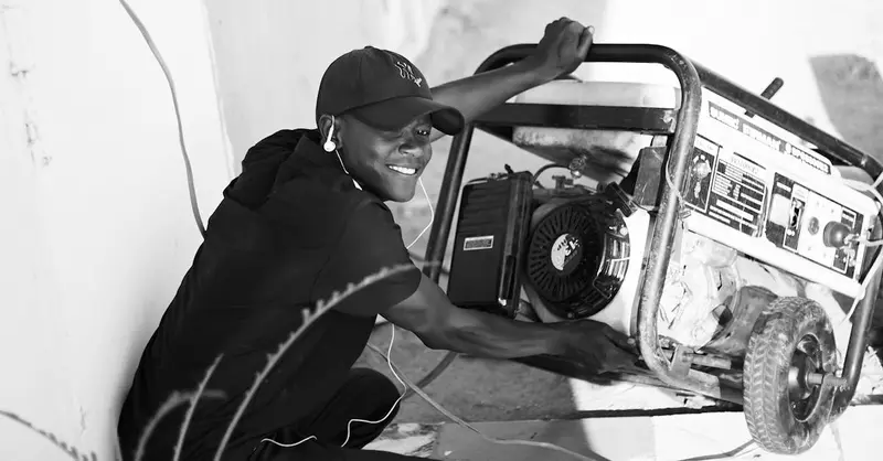 A cheerful young man wearing a cap and earphones operating a portable generator outside.