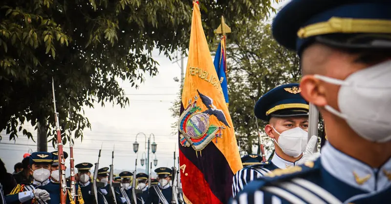 Military personnel in uniforms with masks march in an Ecuadorian parade featuring the national flag.