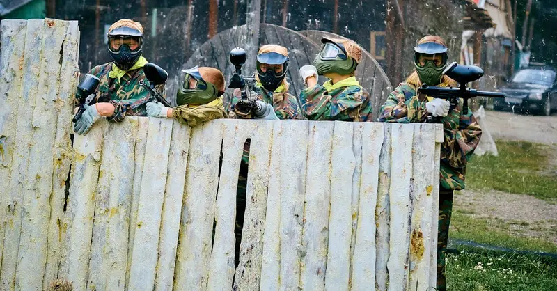 Group of paintball players in camouflage gear aiming over wooden fence outdoors.