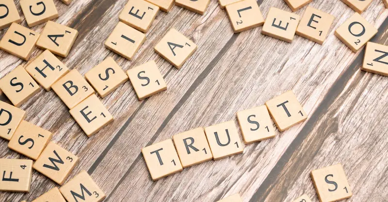Scrabble letter tiles form the word 'Trust' on a wooden table, surrounded by scattered tiles.
