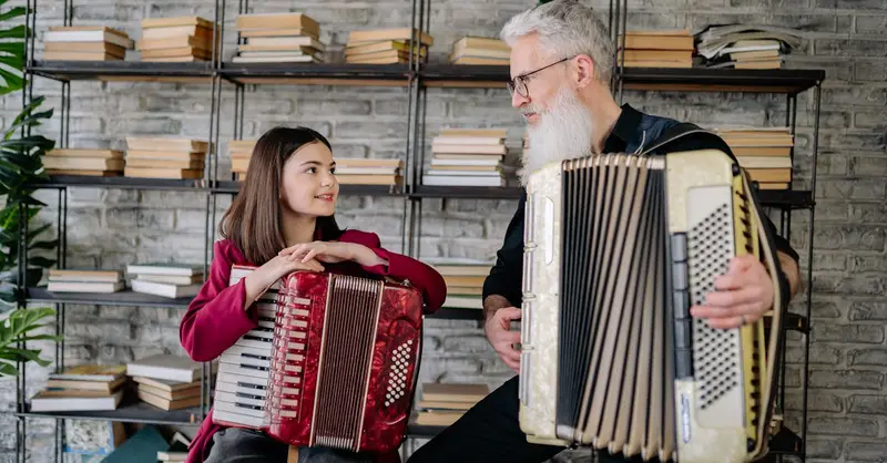 A girl learning accordion from an elderly man in a cozy library setting. Music and education.
