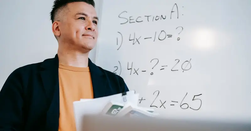 Smiling teacher holding papers, explaining algebra equations on a whiteboard in a classroom setting.