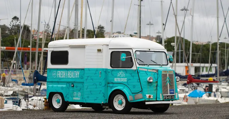 Classic turquoise Citroen van parked at Lisbon marina with boats in the background.