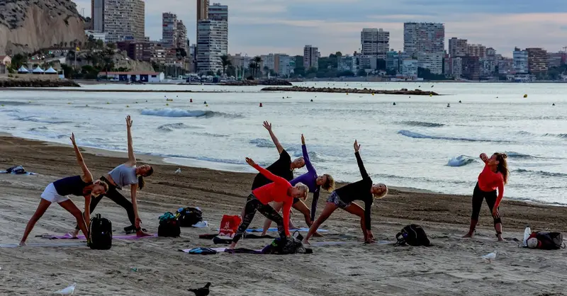 Group practicing yoga on Alicante beach with city skyline backdrop.