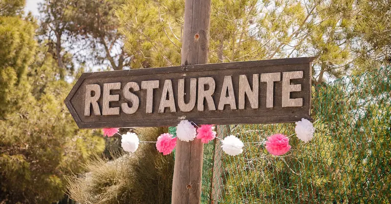 A rustic wooden restaurant sign in Spain, adorned with festive pink and white decorations amidst a natural outdoor setting.