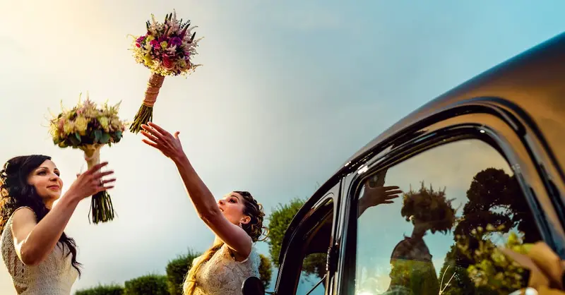 Two brides joyfully exchange bouquets beside a vintage car at their vibrant wedding in Toledo.