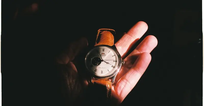 Close-up of a wristwatch with leather strap held in a hand, accented by dramatic lighting.