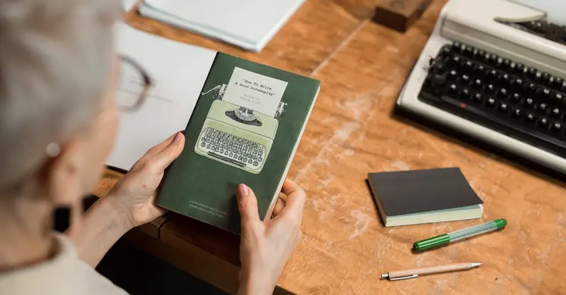 Elderly woman holding a book at a wooden desk with a classic typewriter, creating a retro office vibe.