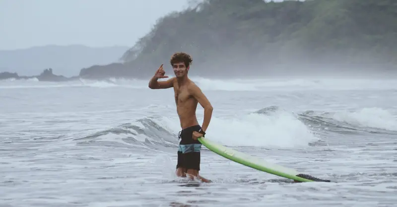 A young surfer stands confidently in ocean waves with his surfboard on a misty day.