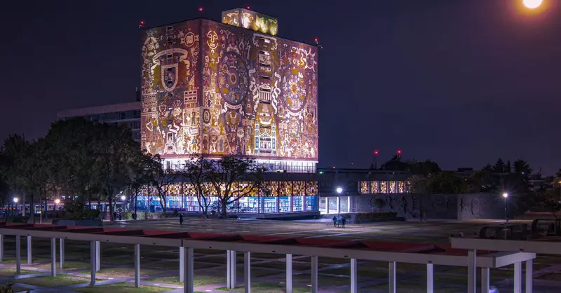 Illuminated UNAM Central Library with vibrant murals and full moon in Mexico City.