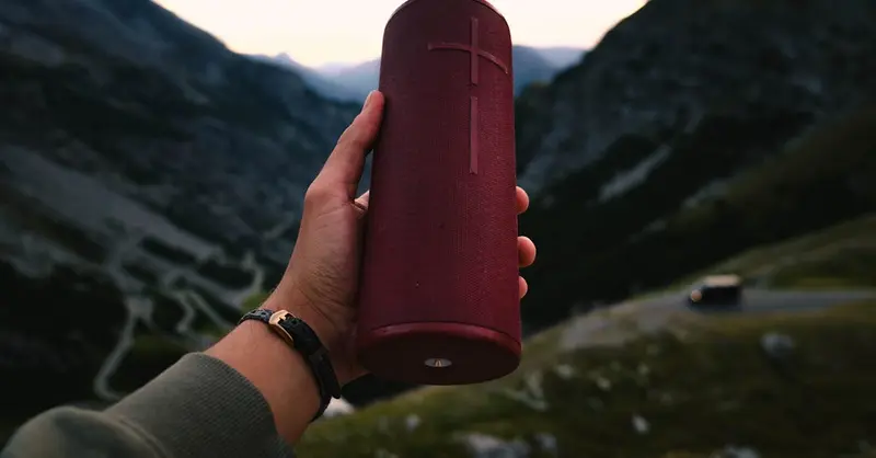 A person holding a red portable speaker with picturesque mountain backdrop.