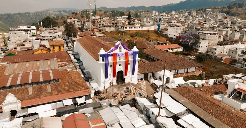 Aerial view of a church decorated with colorful banners amidst a bustling urban town landscape.