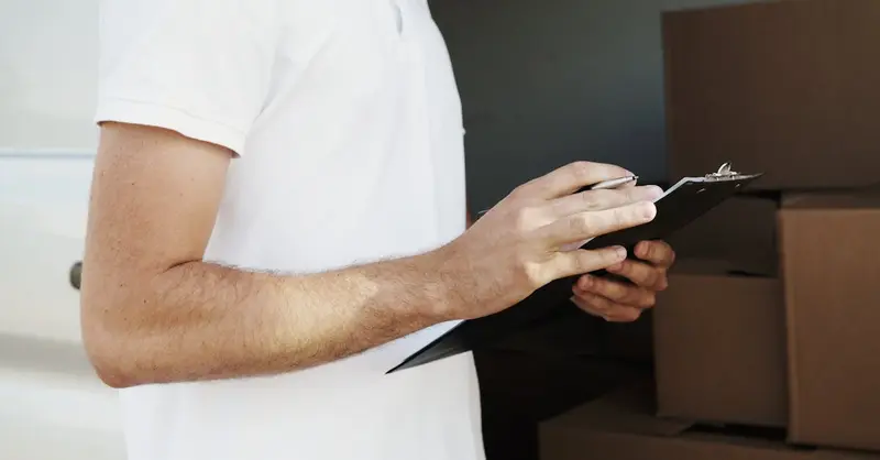 A delivery person in a white shirt holding a clipboard and pen near a vehicle with boxes.