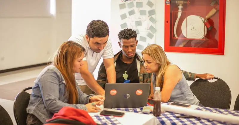 A diverse group of adults collaborating around a laptop during a team meeting in a professional setting.