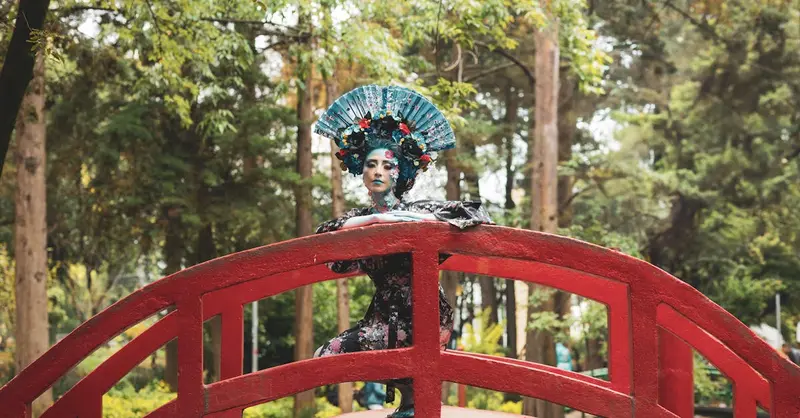 A vibrant performer in floral costume stands on a red bridge amidst a lush park in Mexico City.