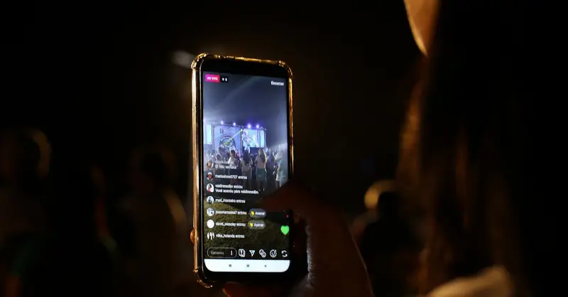 A woman live streams a concert using her smartphone at night. Vibrant stage in view.