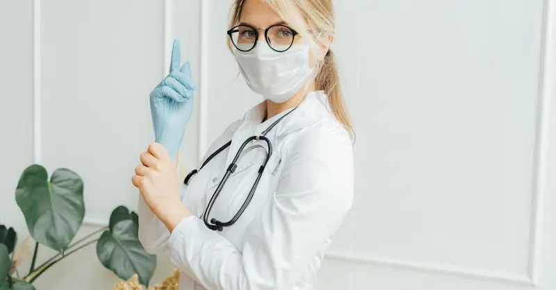 Healthcare professional in a white coat wearing a face mask and gloves, holding a stethoscope.